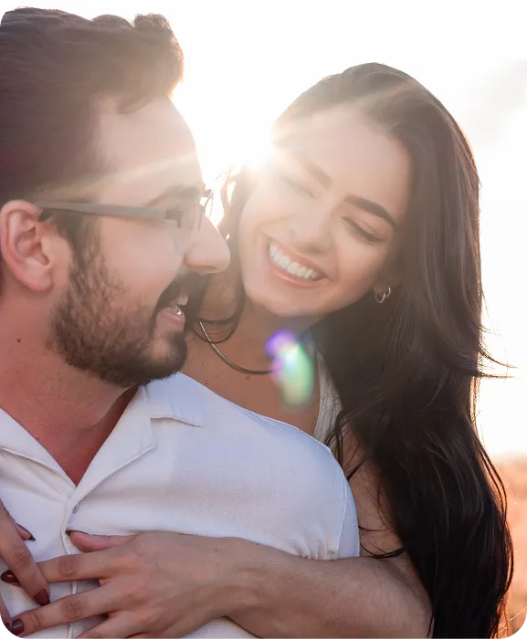 Smiling woman with long dark hair hugging man with glasses and beard in bright sunlight.