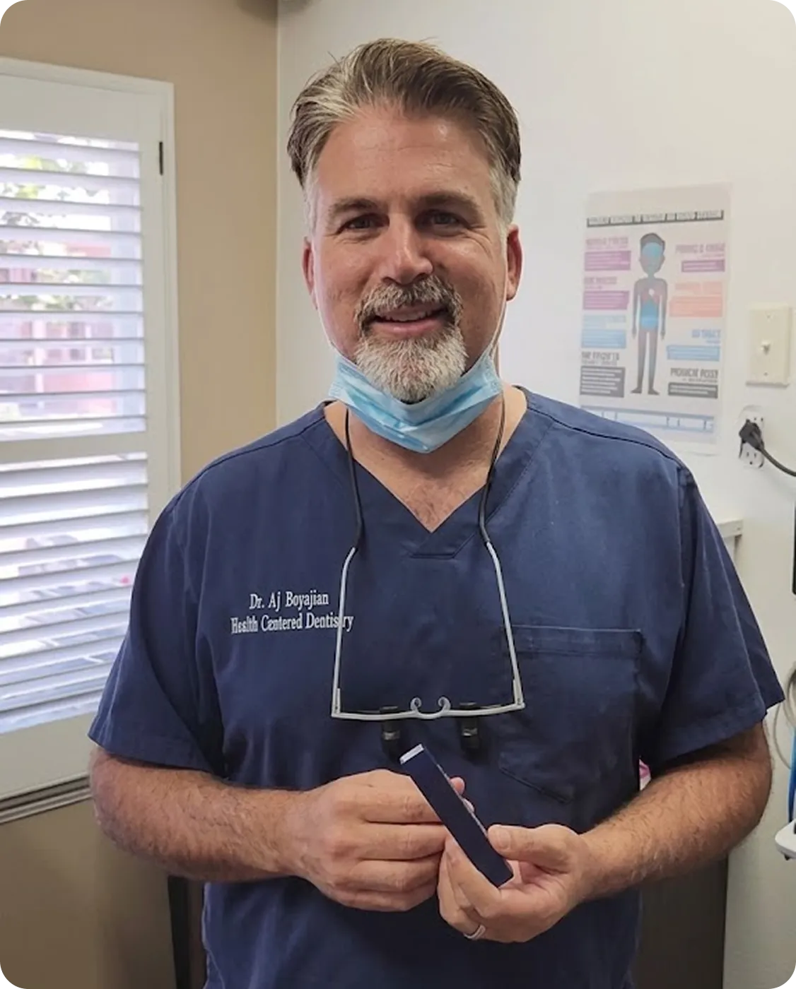 Man in dental scrubs with mask under chin and magnifying glasses around neck holding a small rectangular object.