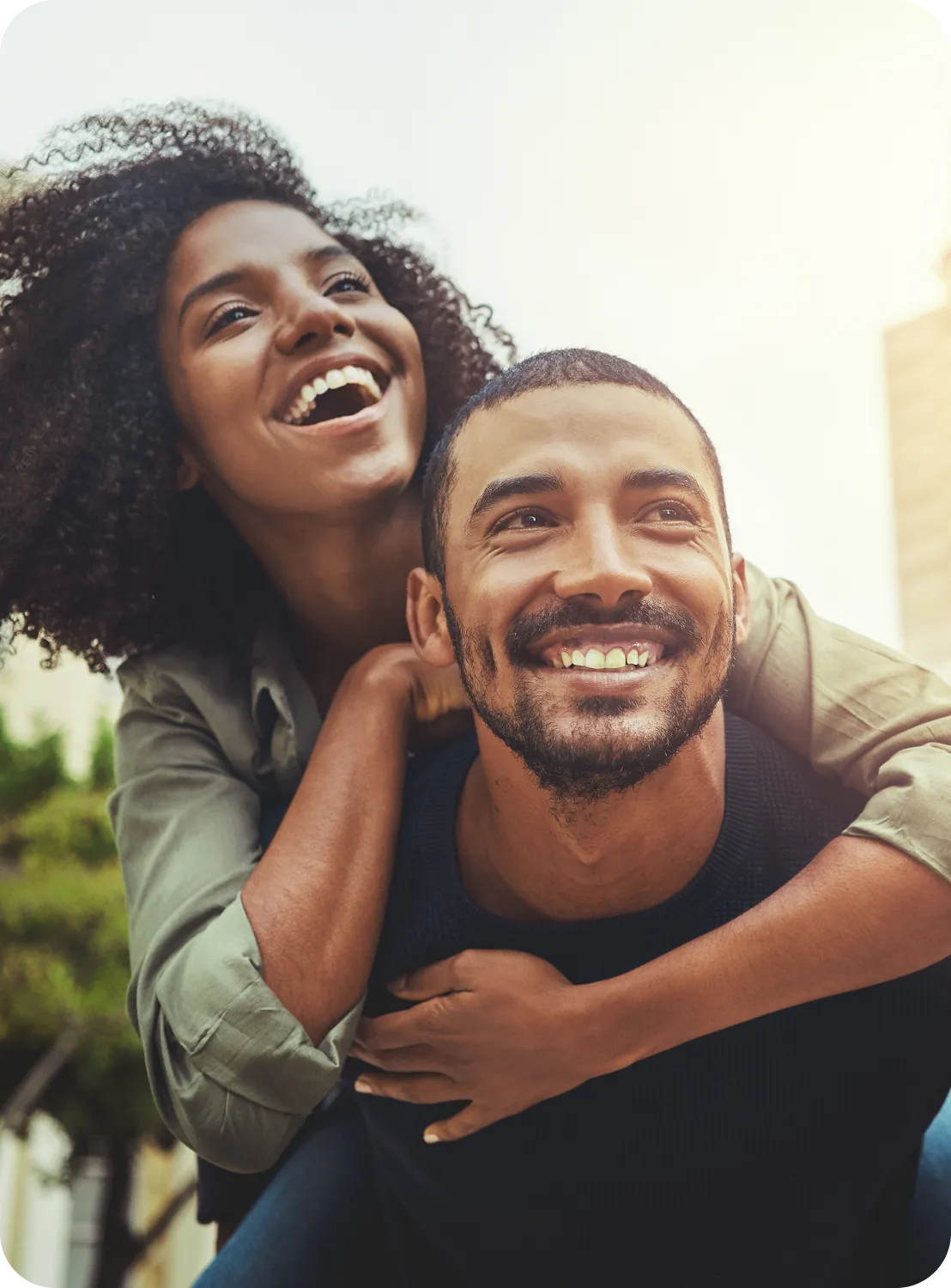 Smiling woman with curly hair piggybacks a smiling man with beard outdoors in daylight.