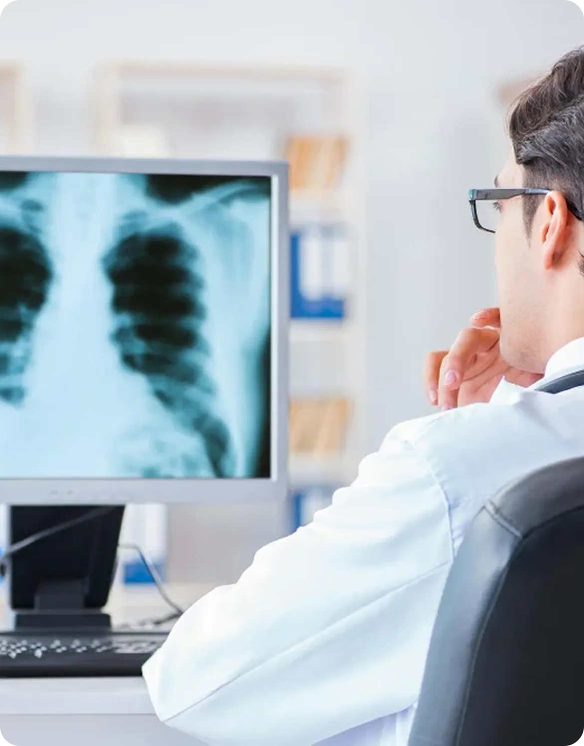 A doctor wearing glasses and a white coat is seated and attentively examining a chest X-ray displayed on a computer monitor in a medical office.