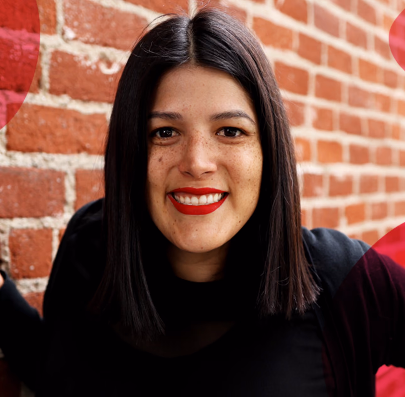 Smiling woman with straight black hair and red lipstick against a red brick wall background.