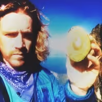 Man with long hair and blue jacket holding a halved avocado in outdoor mountainous setting.