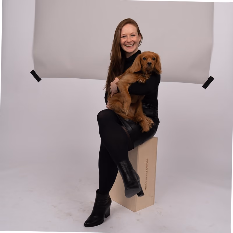 Smiling woman in black holds a brown dog while sitting on a wooden block in a studio.