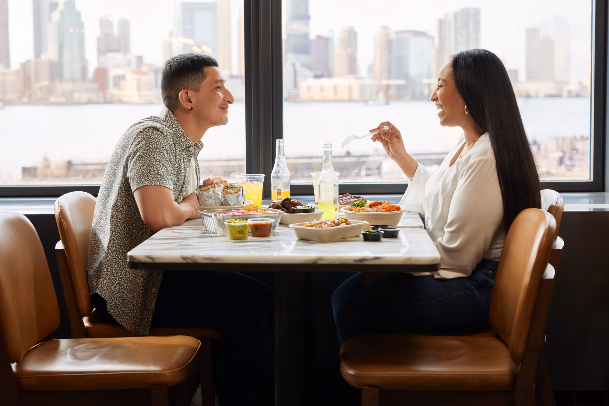 A man and woman sitting at a table by a window, smiling and sharing a meal with city skyline behind them.