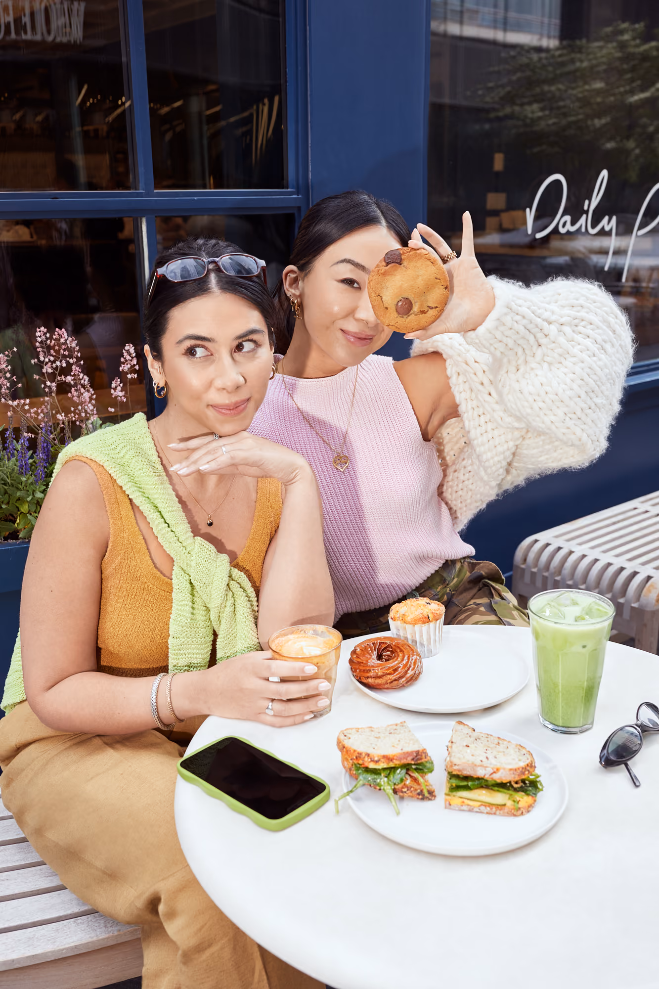 Two women enjoying coffee, sandwiches, and pastries at an outdoor café table.