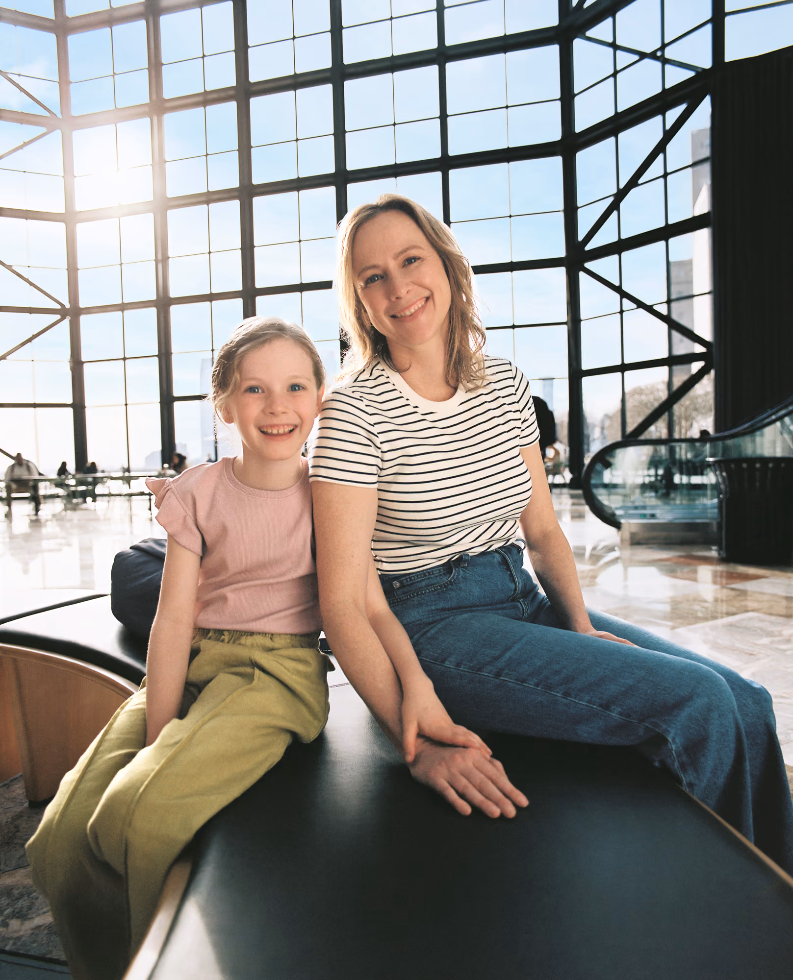 Smiling woman and young girl sitting side by side on a bench inside a brightly lit building.