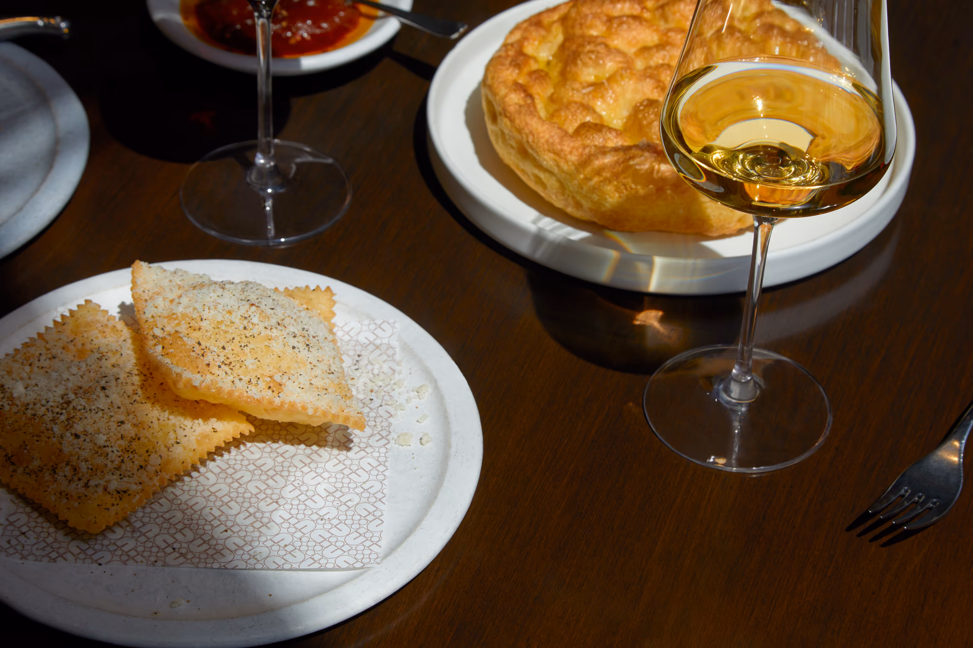 Two square crackers with seasoning on a plate, a round bread, and a glass of white wine on a table.