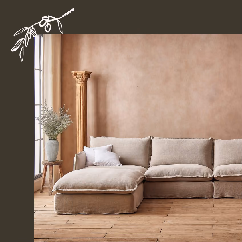 Beige sectional sofa with cushions in front of a brown wall, near a window with a vase on a stool.