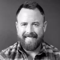 Smiling man with beard wearing a plaid shirt against a plain background.