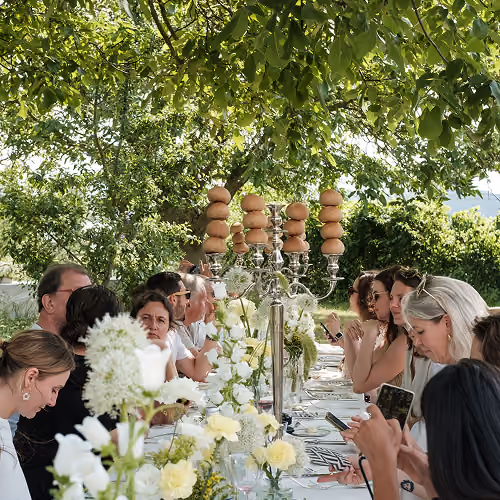 Personnes assises autour d'une longue table décorée de fleurs blanches sous un grand arbre vert.