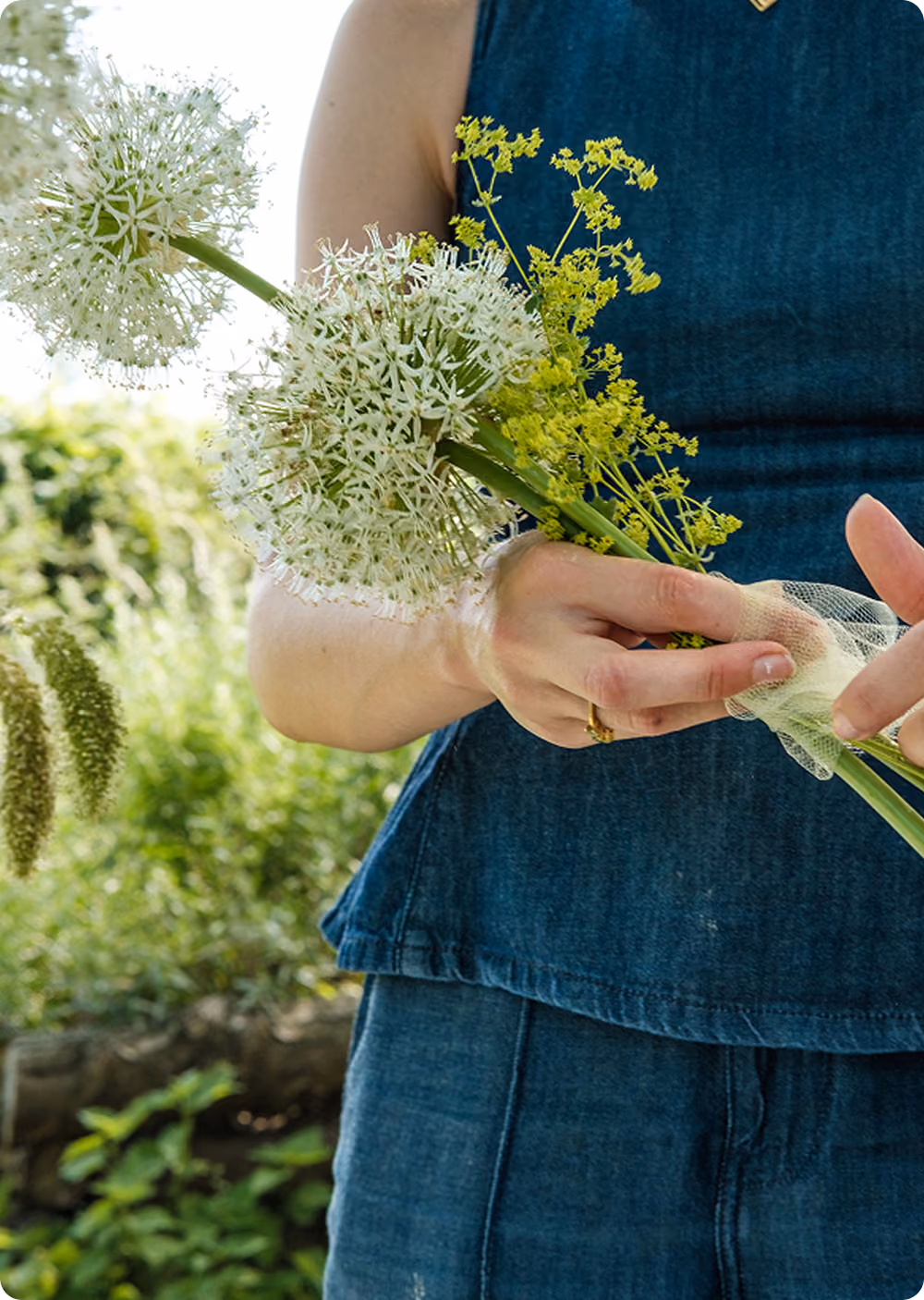 Person in blue denim dress holding white and yellow flowering stems outdoors.