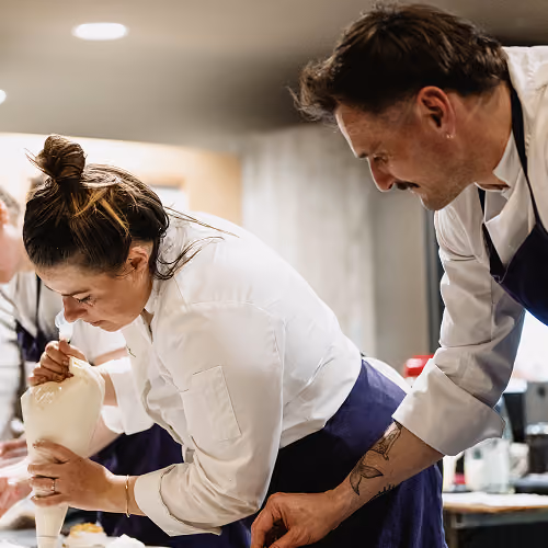 Deux chefs en tenue blanche et tabliers observent et travaillent sur une préparation culinaire.