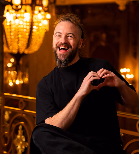 Un homme souriant forme un cœur avec ses mains dans un intérieur luxueux et éclairé par des chandeliers.