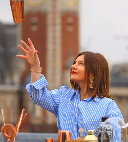 Femme en chemise rayée bleue attrapant un verre en cuivre avec des accessoires de bar flous.