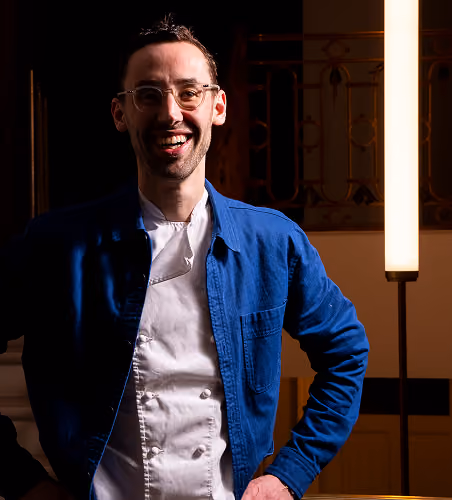 Homme souriant portant une chemise bleue et une veste blanche dans un intérieur sombre.