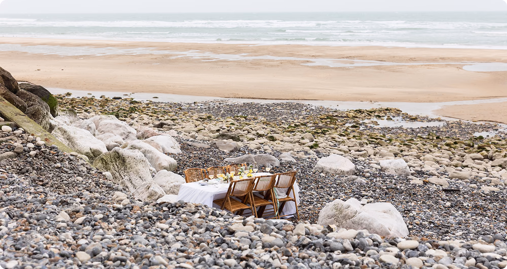 Table set with chairs on rocky shore near sandy beach and ocean waves in the distance.