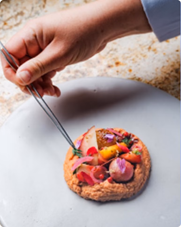 Main utilisant une pince pour décorer un dessert gourmet avec des fruits et des fleurs comestibles.