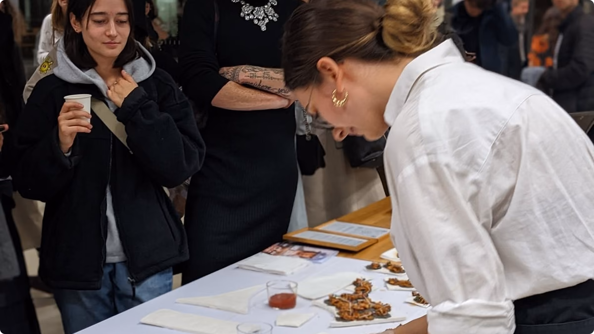 Femme avec chemise blanche préparant des amuse-gueules sur une table devant des observateurs.