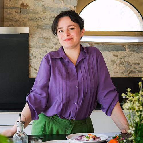 Jeune femme souriante en chemise violette et pantalon vert debout dans une cuisine près d'une table avec des plats.