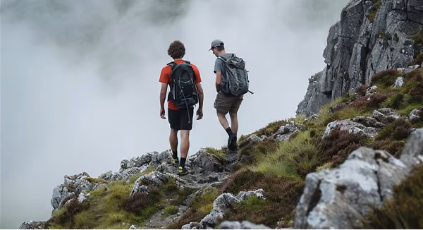Two hikers with backpacks walking on a rocky mountain trail surrounded by fog and grass.