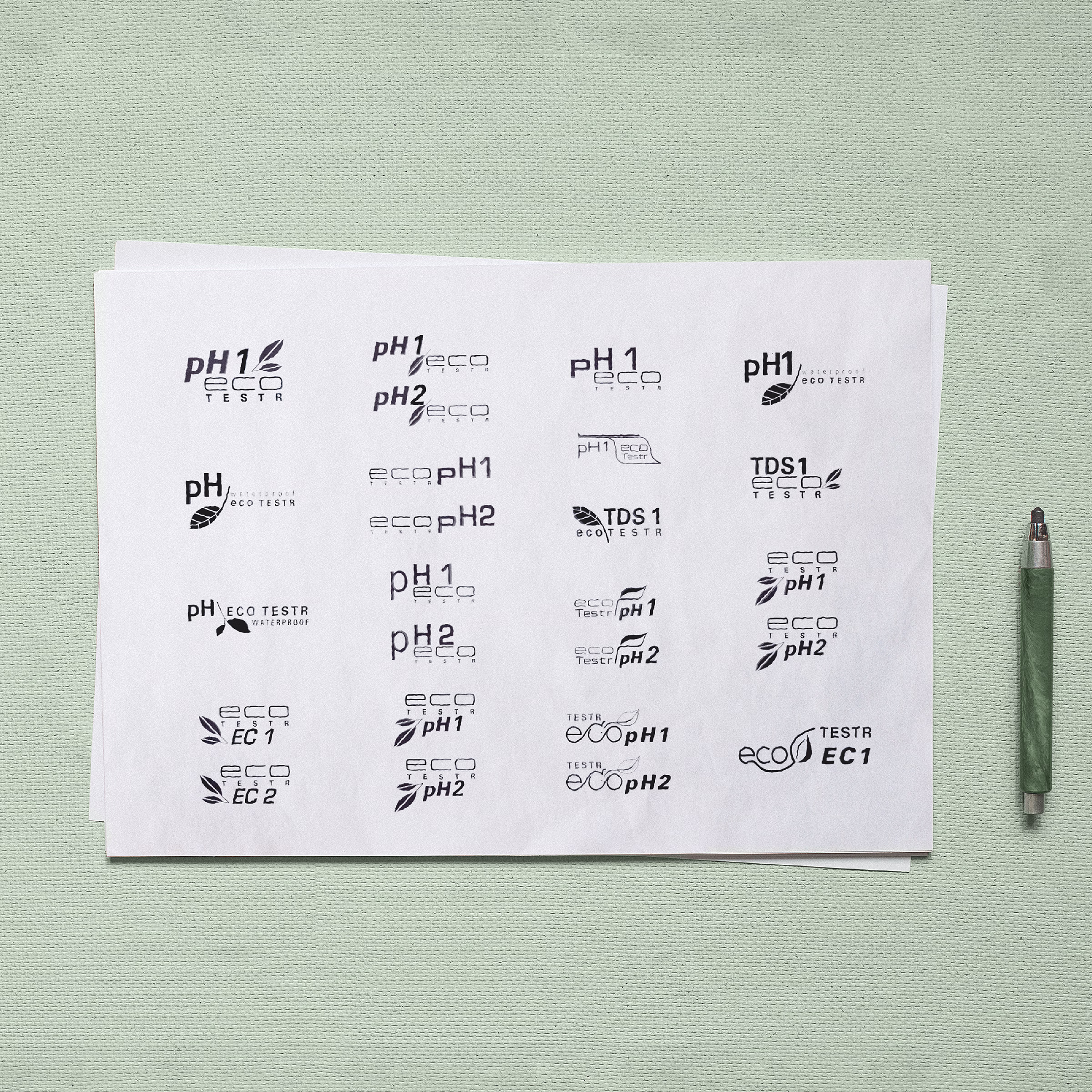 Printed sheet of EcoTestr logo explorations photographed on a green textile background, showing more than twenty mark directions combining pH, EC, TDS, and Salt product variants with leaf motifs, ECO wordmarks, and waterproof callouts.