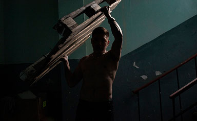 Silhouetted shirtless man carrying wooden ladder on his shoulder in dim stairwell.