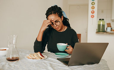 Person wearing glasses holding a green cup and rubbing forehead at a table with laptop and snacks.