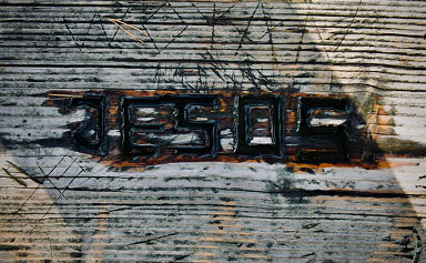 Rusty metal letters spelling 'JESUS' on worn wooden slats with nail markings.