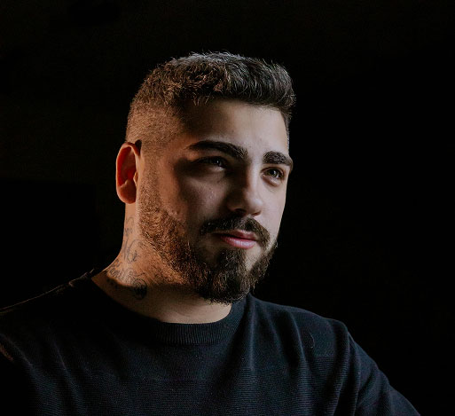 Man with short dark hair, beard, neck tattoos, and dark sweater looking slightly to the side in low light.