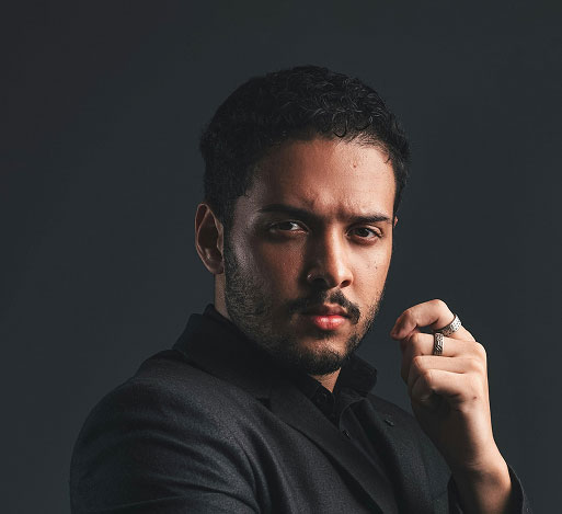 Man with dark hair and beard wearing a black suit, posing with fingers near face against dark background.