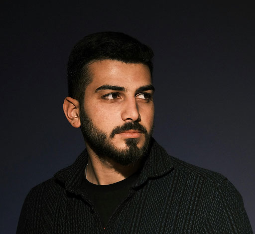 Bearded man with dark hair wearing a dark textured jacket looking to his left against black background.