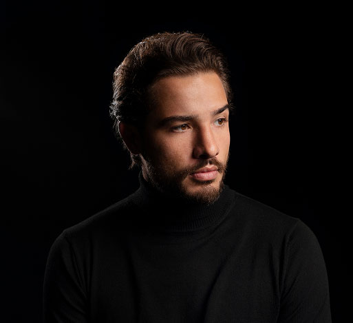 Young man with beard wearing a black turtleneck looking thoughtfully to the side on a black background.