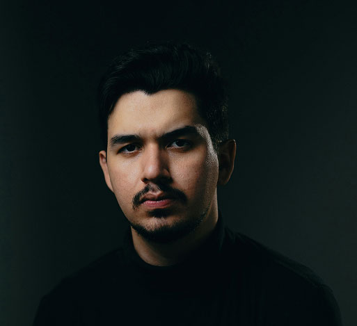 Serious young man with dark hair and mustache wearing a black turtleneck against a dark background.