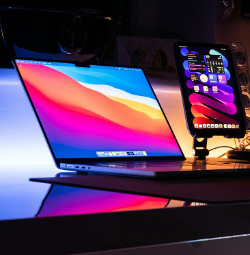 Laptop and smartphone side by side on a desk with colorful screen reflections in a dimly lit room.
