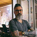 Bearded man with glasses and tattoos crossing arms, standing indoors by a window.