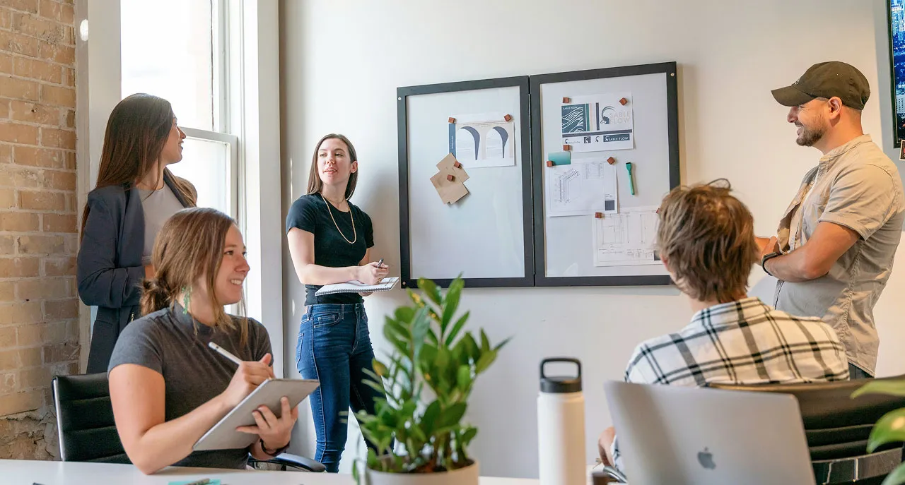 Five people in a meeting room, two women presenting ideas on a whiteboard to seated colleagues.