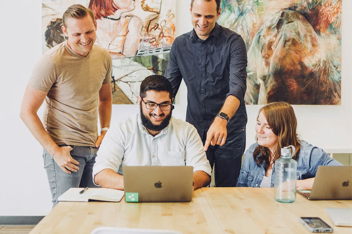 Four coworkers smiling while looking at a laptop around a table with notebooks and water bottle.