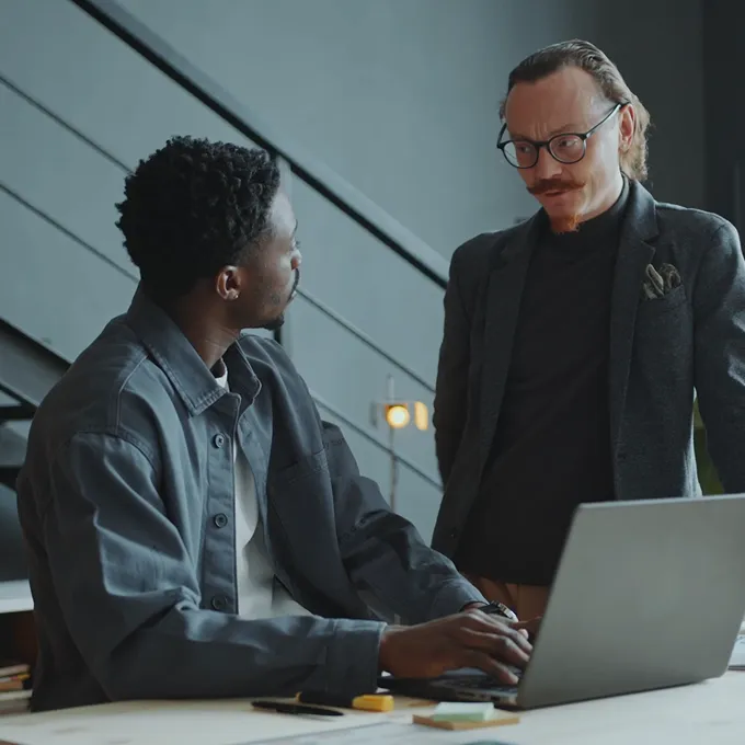 Two men at a desk with a laptop, one sitting and typing, the other standing and watching.