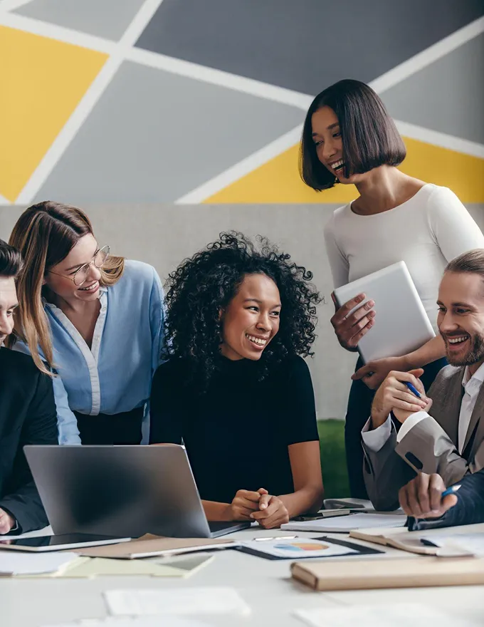 Five diverse coworkers smiling and discussing around a laptop and papers in a modern office.