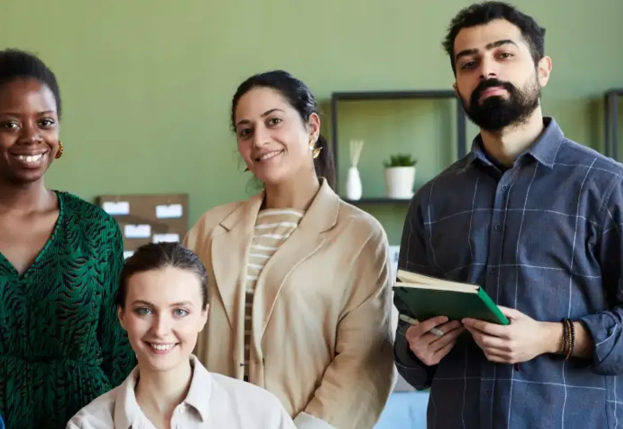 Four diverse professionals smiling and posing in a modern office with books and decor in the background.