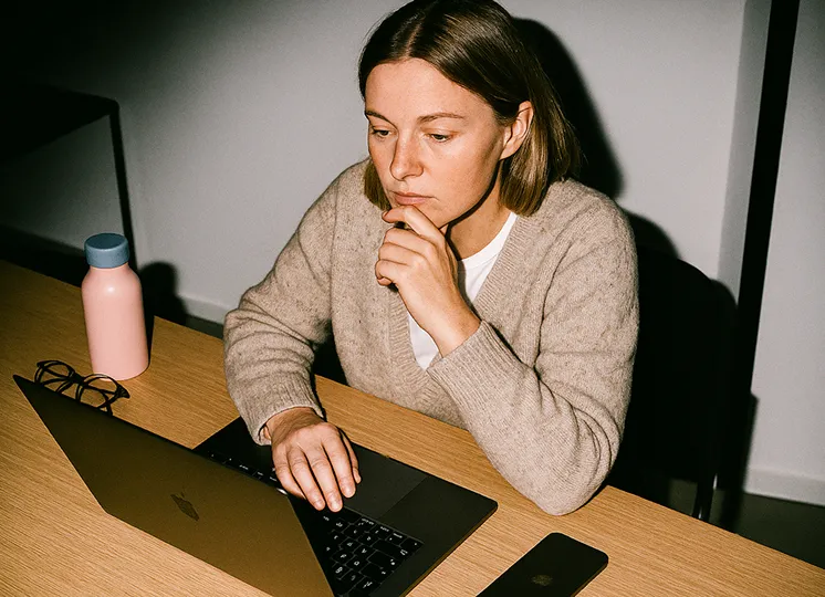 Woman in a beige sweater focused on laptop at a wooden desk with glasses, water bottle, and phone nearby.