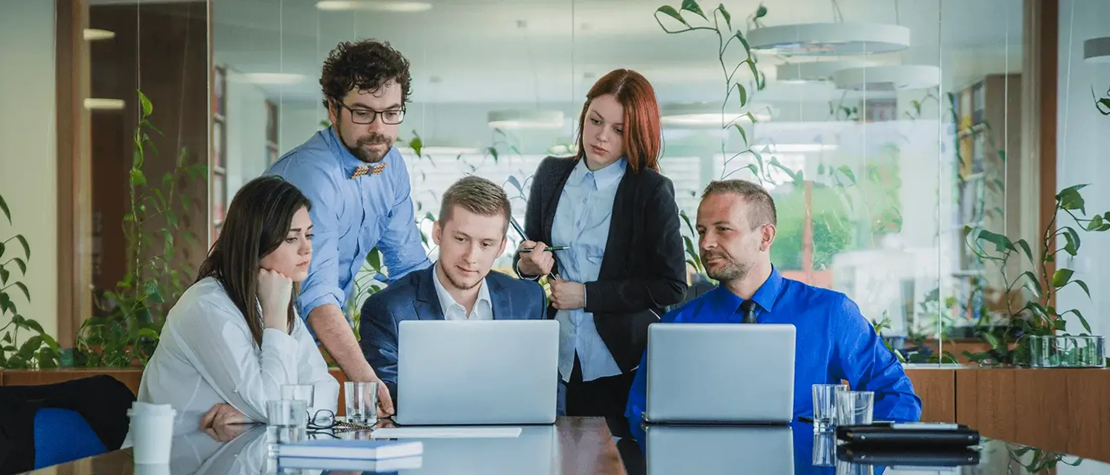 Five business colleagues gathered around a table, two using laptops, discussing work in an office.