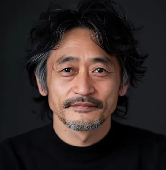 Middle-aged man with black and gray hair and a short beard wearing a black sweater against a dark background.