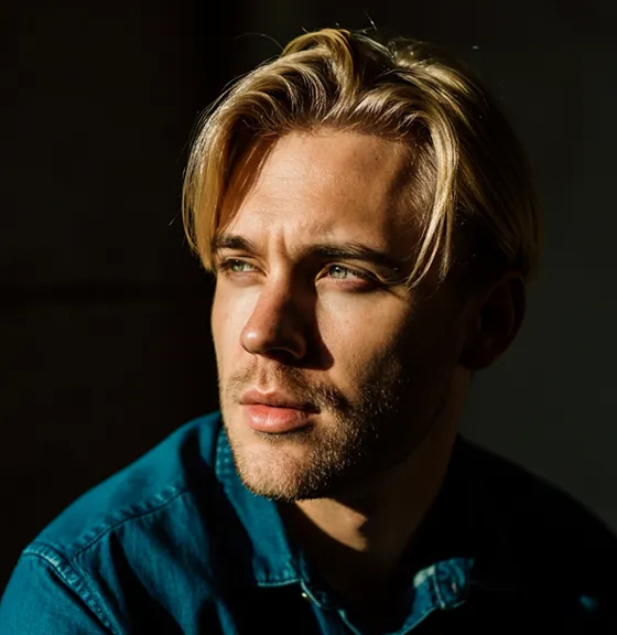 Portrait of young blond man in teal shirt looking thoughtfully to the left in low light.