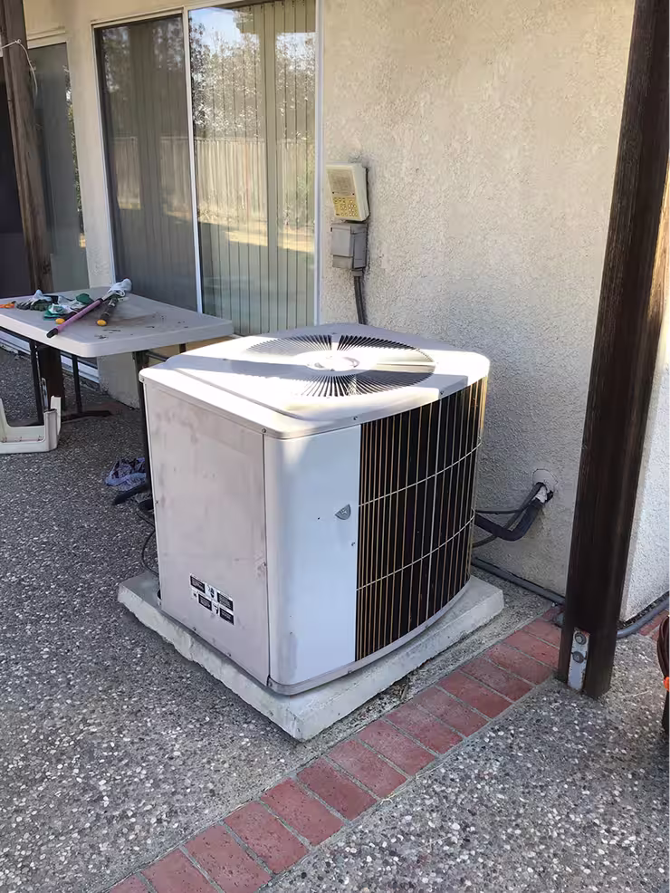 Outdoor air conditioning unit on a concrete base next to a beige stucco wall with a sliding glass door and a folding table holding garden tools and gloves.
