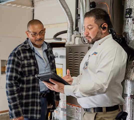 Technician in white shirt showing information on a tablet to a man in a plaid shirt inside a utility room with HVAC equipment.