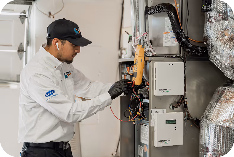 Technician in white shirt and black cap working on a furnace with a multimeter in a residential garage.