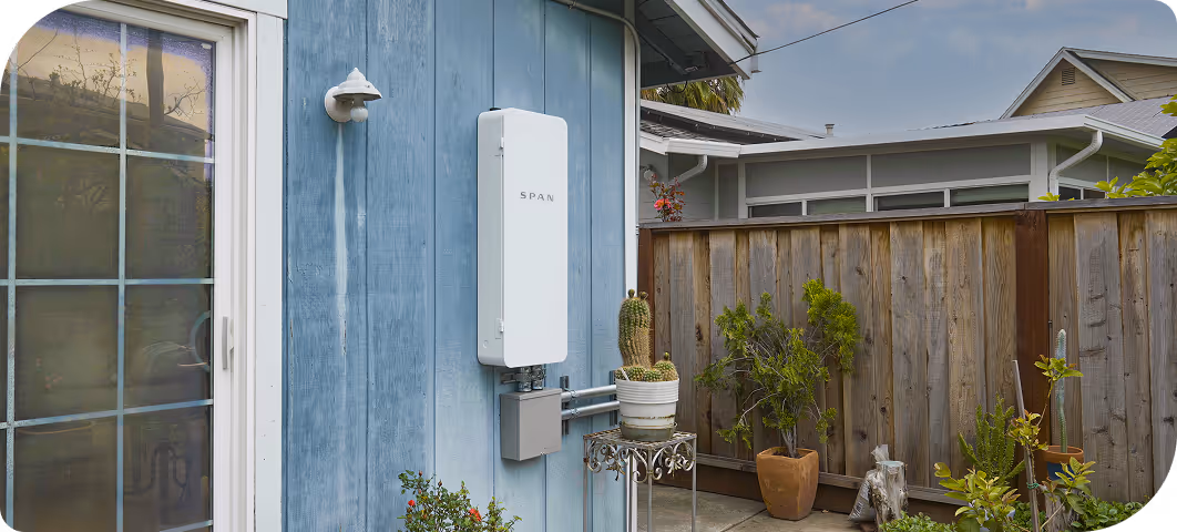 Smart home electrical panel labeled SPAN mounted on the exterior blue wall of a house near potted plants and a wooden fence.