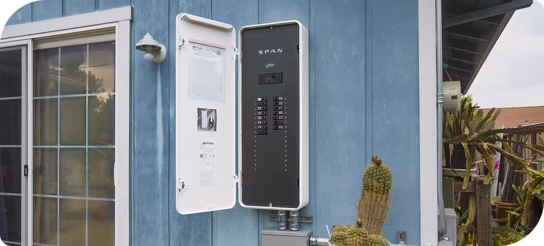 Open SPAN electrical panel mounted on the exterior blue wall of a house near a sliding glass door and a cactus plant.