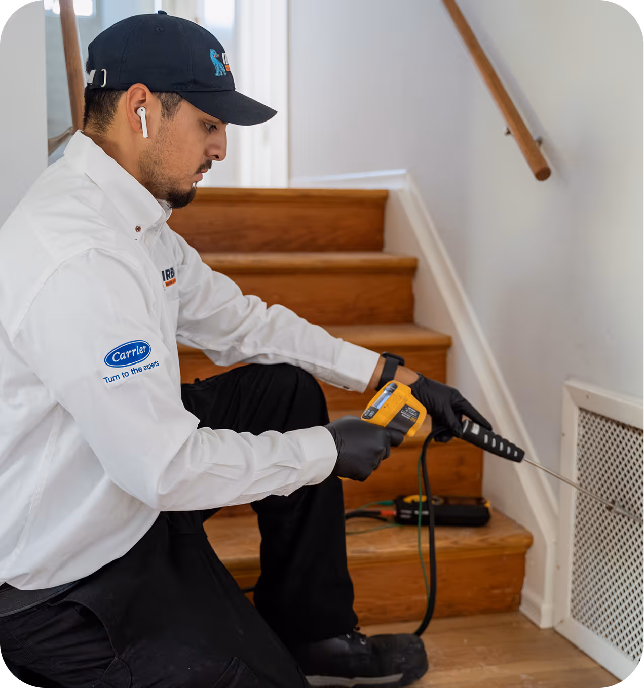 Technician in white shirt and black cap using a temperature gun to inspect a vent near wooden stairs.